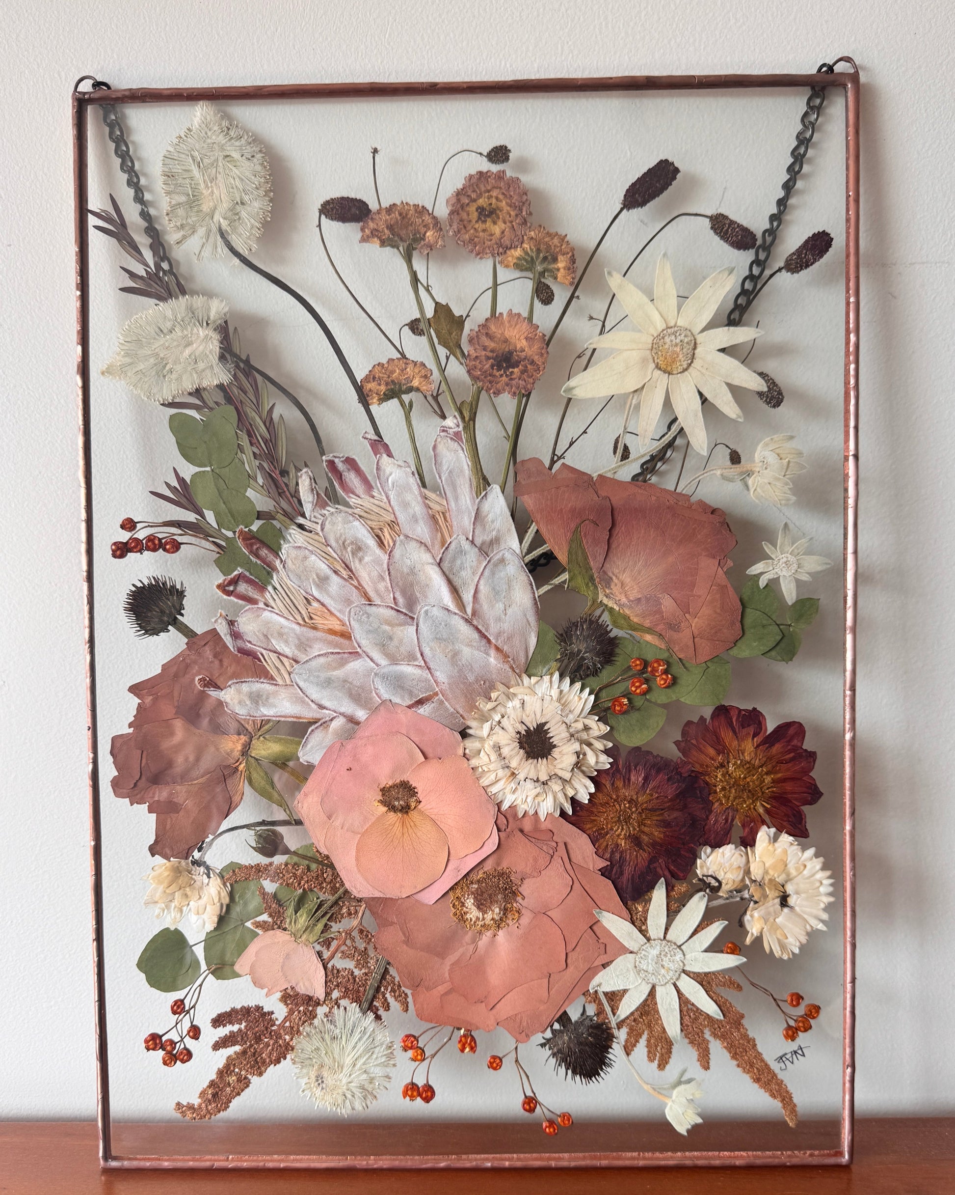 Pressed flower with King protea framed in a copper soldered metal frame on a wooden surface with a white wall background