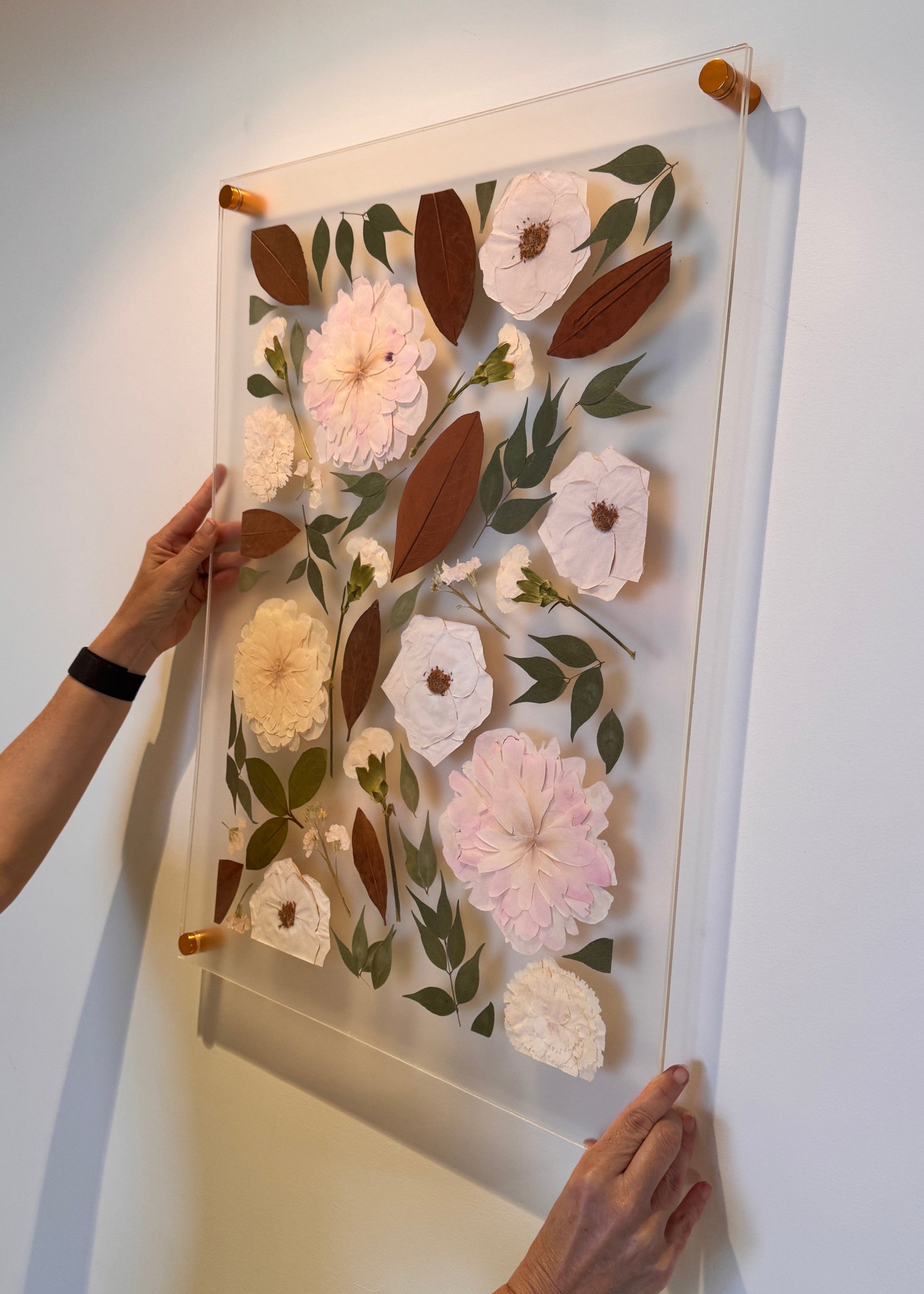 Pressed flower artwork in a double glass frame held by a person against a white wall