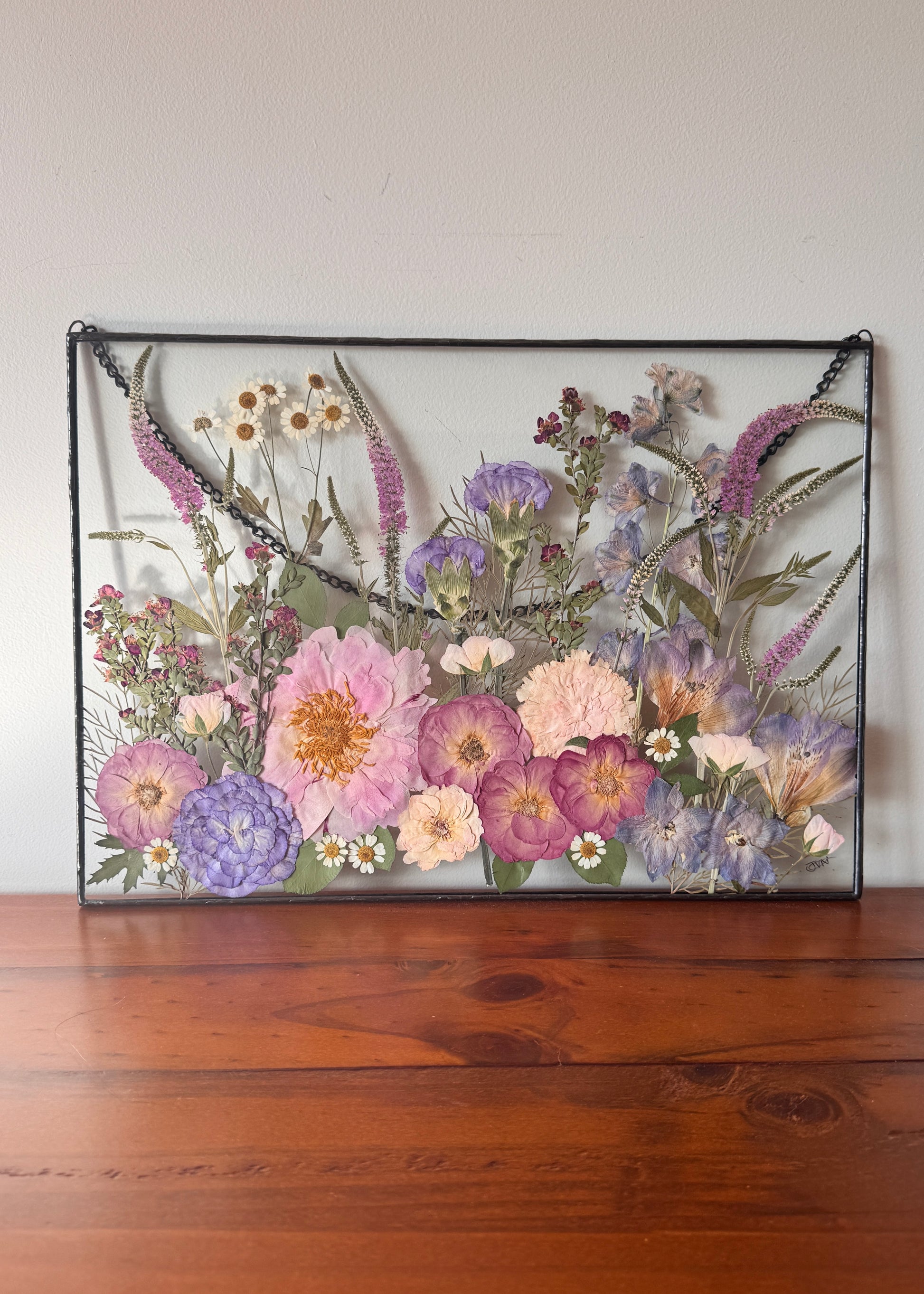 Pressed flower arrangement in a black hand soldered frame on a wooden surface with a white wall background