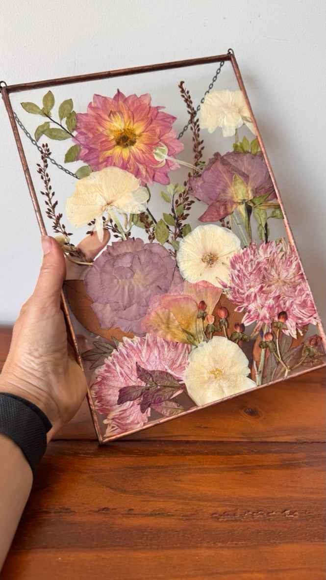 A hand holds a rectangular framed piece of pressed dried flowers with pink, cream, and purple blooms over a wooden table against a light wall.