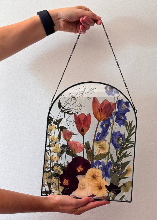 A handcrafted metal arch-shaped frame with a bouquet of preserved flowers behind glass, held by a person against a white background.