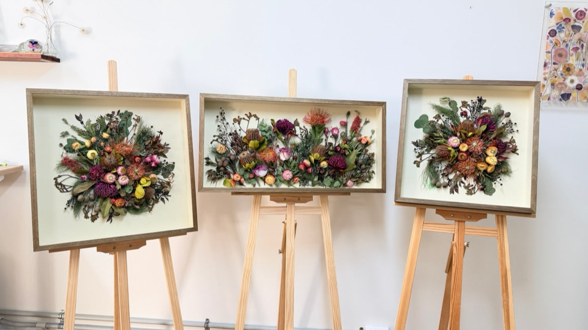 Three framed dried native Australian flower arrangements displayed on wooden easels against a white wall in Created with Joy Arts  indoor studio