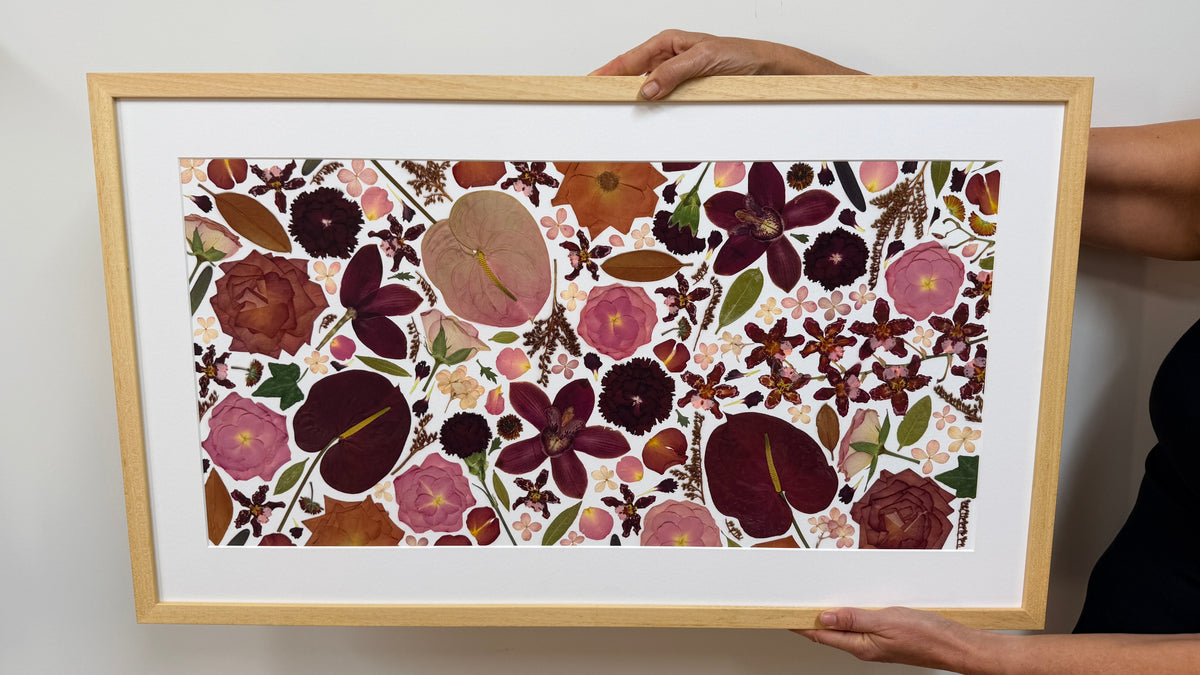 A framed floral art print held by a person's hands against a plain light gray wall. The artwork features a dense arrangement of pressed flowers and botanical elements in pink, burgundy, brown, orange, and green on a white background, enclosed in a light wood frame with a wide white mat.