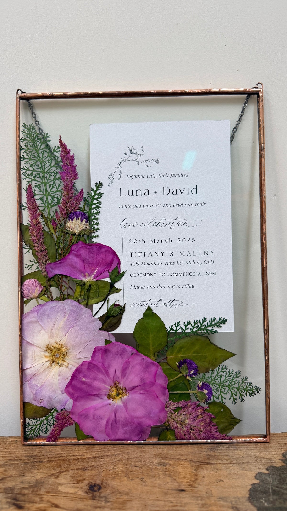 A framed floral wedding invitation card with purple and pink flowers and green foliage inside a copper soldered metal frame. 