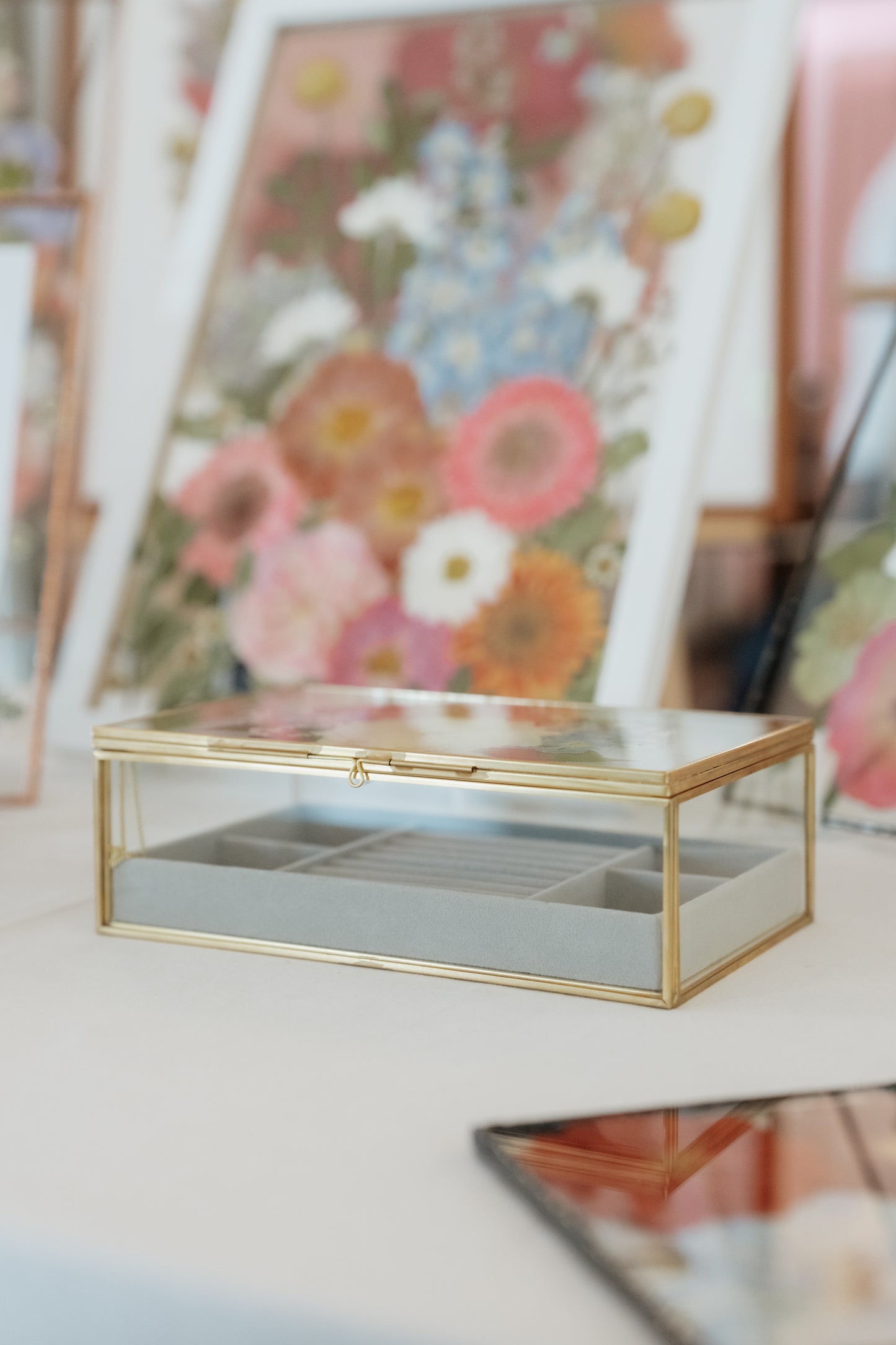 A rectangular glass jewelry box with a gold metal frame and a light gray interior organizer tray, set on a white surface with a blurred floral background.