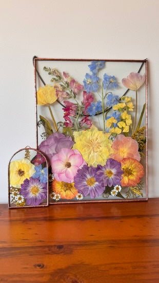 A framed pressed flower arrangement with a copper-toned metal frame, displayed on a wooden surface against a plain light wall. A smaller arch matching frame sits beside it.