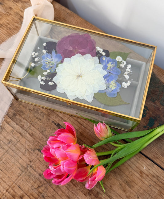 A rectangular glass keepsake box with a gold frame, containing pressed flowers including a large white flower, blue flowers, and a purple flower, displayed on a rustic wooden table next to pink tulips.