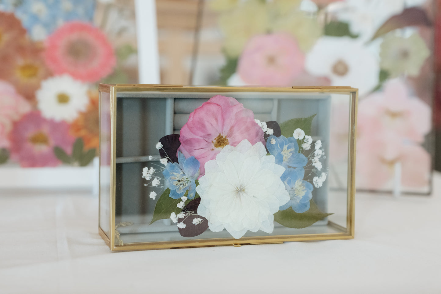 A rectangular glass shadow box with a gold metal frame, containing pressed flowers including a large white flower, a pink flower, and blue baby's breath, displayed on a white surface with a blurred floral background.