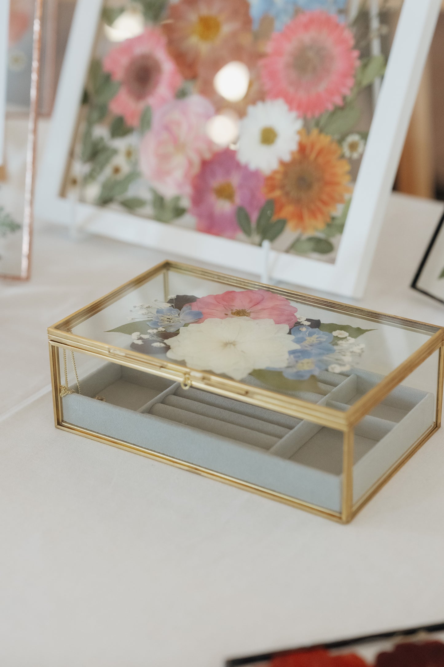 A rectangular glass jewelry box with a gold metal frame and a lid decorated with pressed flowers, resting on a white surface with a blurred floral picture frame in the background.
