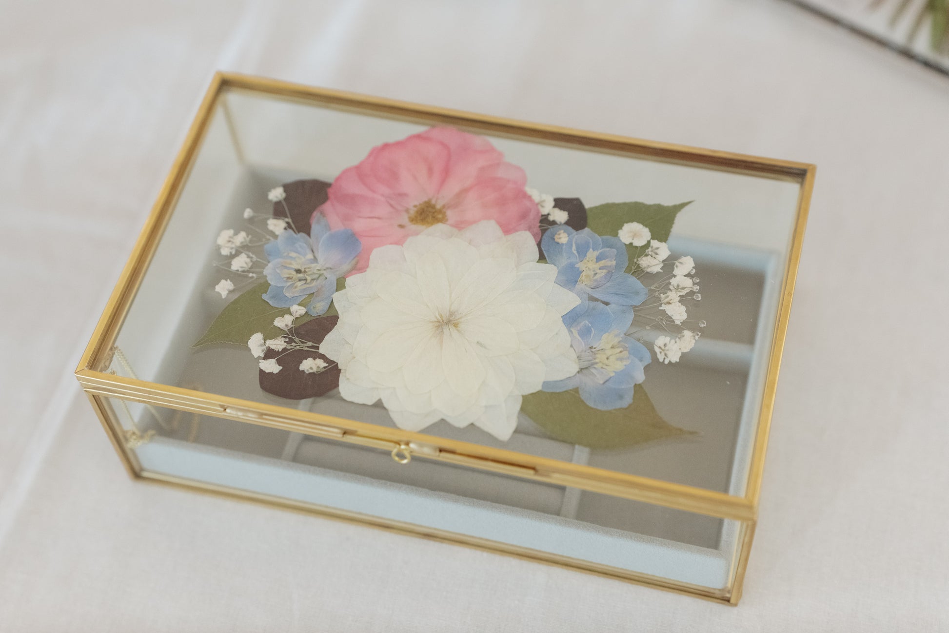 A rectangular glass jewelry box with a gold metal frame, featuring a pressed floral design on the lid with a large white flower, pink flower, blue flowers, and green leaves, set against a white fabric background.